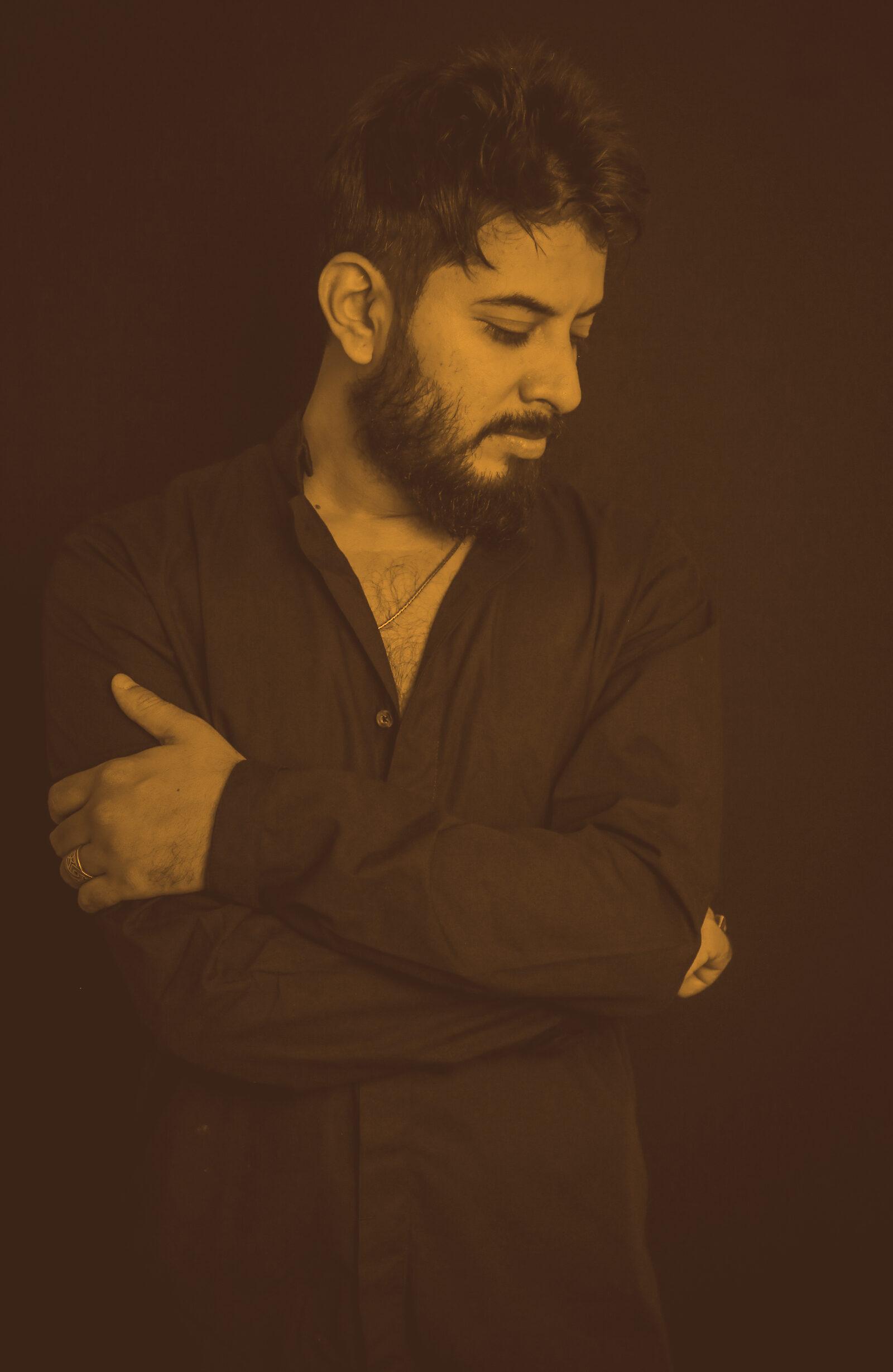 Abhi Maisekar with arms folded, looking down, against a black background.